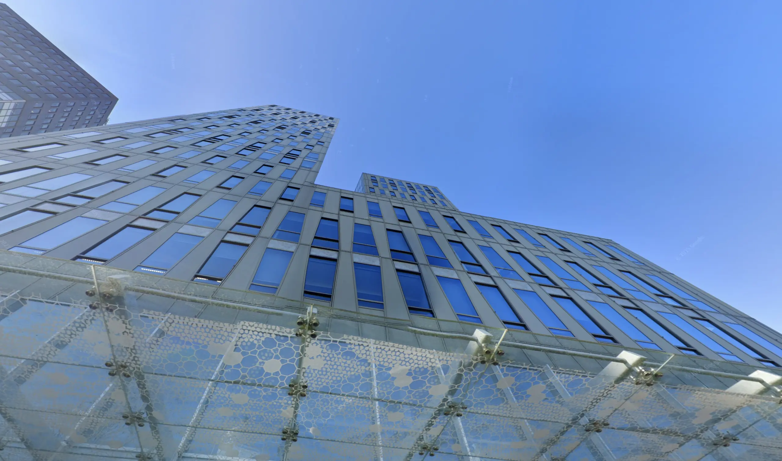 Tall modern building with glass windows and a decorative patterned canopy, viewed from a low angle. The clear blue sky adds to the structure's sleek appearance.
